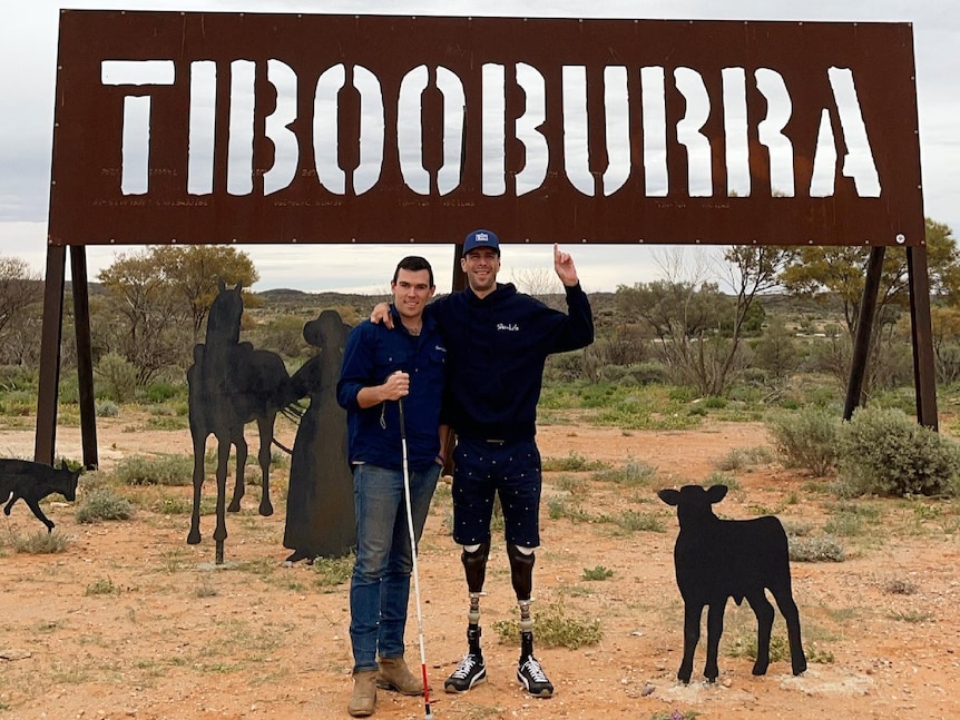 Two men, one with prosthetic legs showing, smile for the camera, with a goat, in front of the Tibooburra town sign. Ausnew Home Care, NDIS registered provider, disability