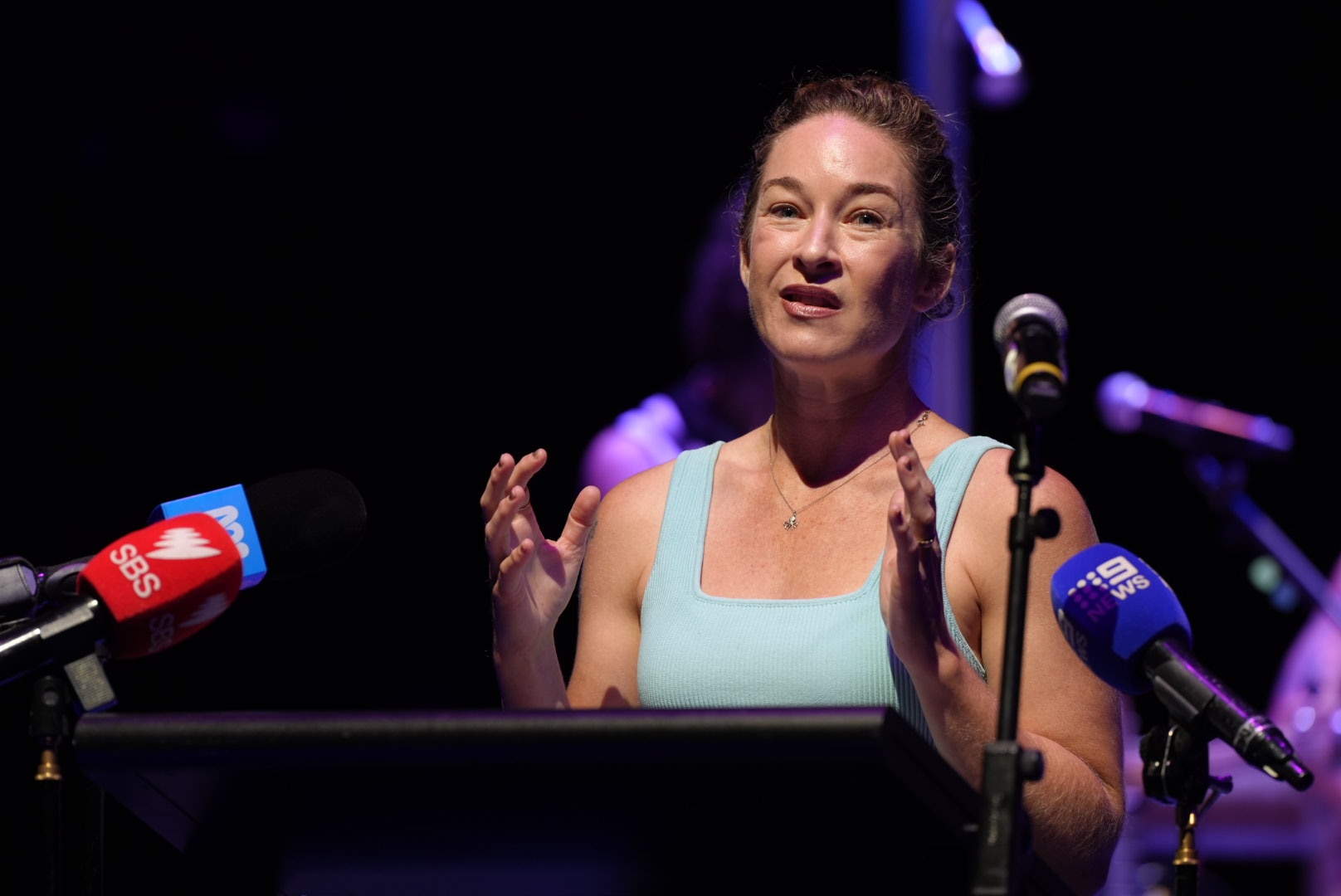Una mujer con un top azul claro y el pelo recogido habla ante los micrófonos de los medios de comunicación en un teatro.