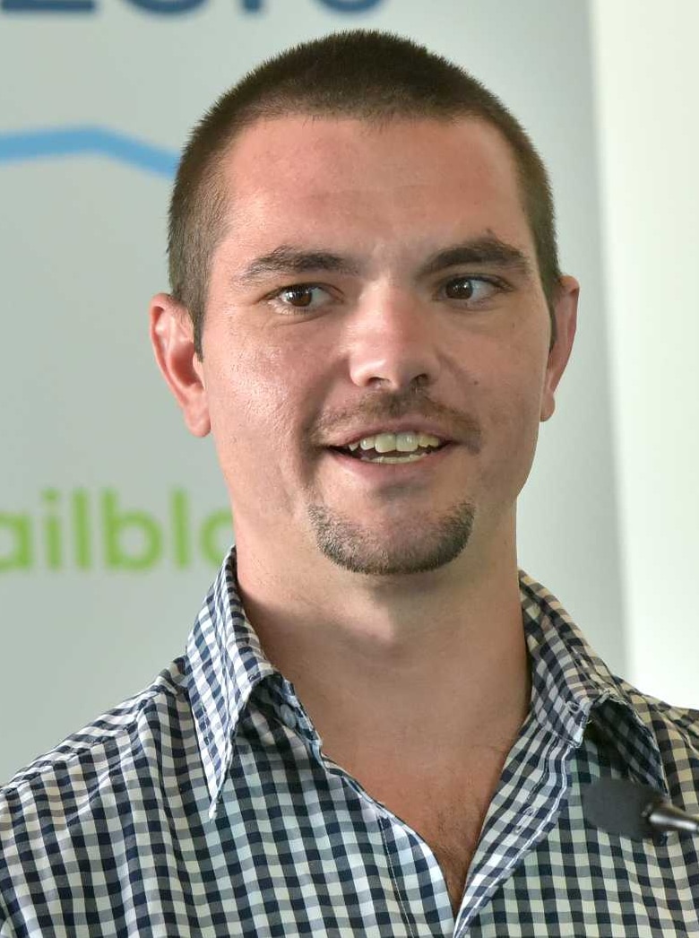 A young man with short brown hair and a check shirt stands in front of a lectern.