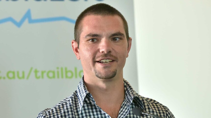 A young man with short brown hair and a check shirt stands in front of a lectern.