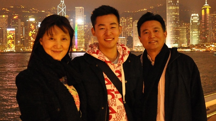 A young man stands in the middle of his mum and dad at night time, with a city skyline lit up behind them.