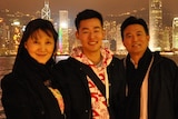 A young man stands in the middle of his mum and dad at night time, with a city skyline lit up behind them.