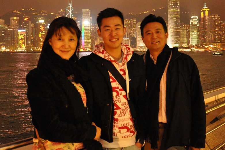 A young man stands in the middle of his mum and dad at night time, with a city skyline lit up behind them.