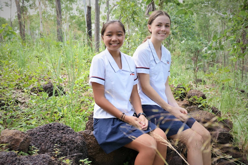 Two teenage girls wearing school uniforms sitting on rocks in the bush.