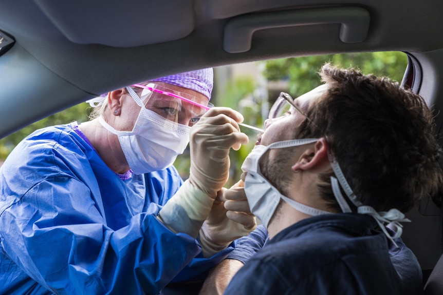 A healthcare worker in full PPE performing a nasal swab on a man in a drive-through COVID testing clinic. 