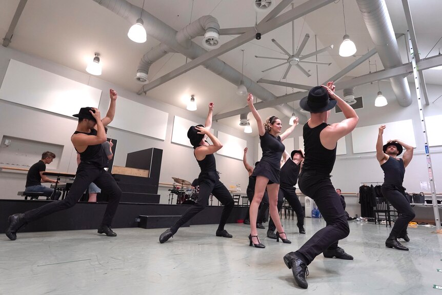 A blonde white woman in black rehearsal clothes and chorus shoes is in the centre of a dance studio surrounded by men in black.