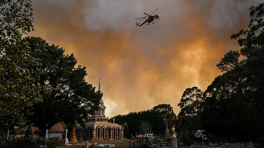 A helicopter is seen in the sky to the right with a monastery underneath it. The sky is orange and grey.
