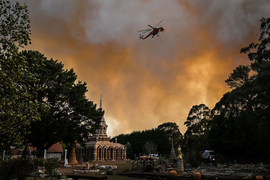 A helicopter is seen in the sky to the right with a monastery underneath it. The sky is orange and grey.