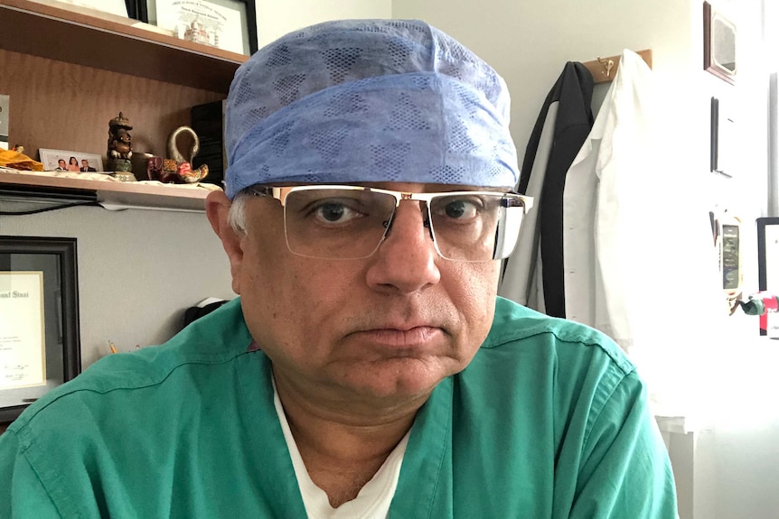 A man wearing scrubs, a doctor's cap and glasses sits in a doctor's office.