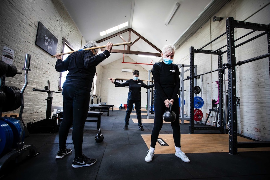 Three women in black clothes do exercises in a gym while wearing face masks.