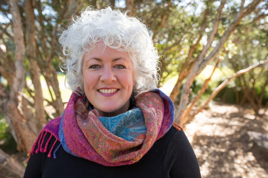 A woman with white curly hair smiles with a tree in the background.