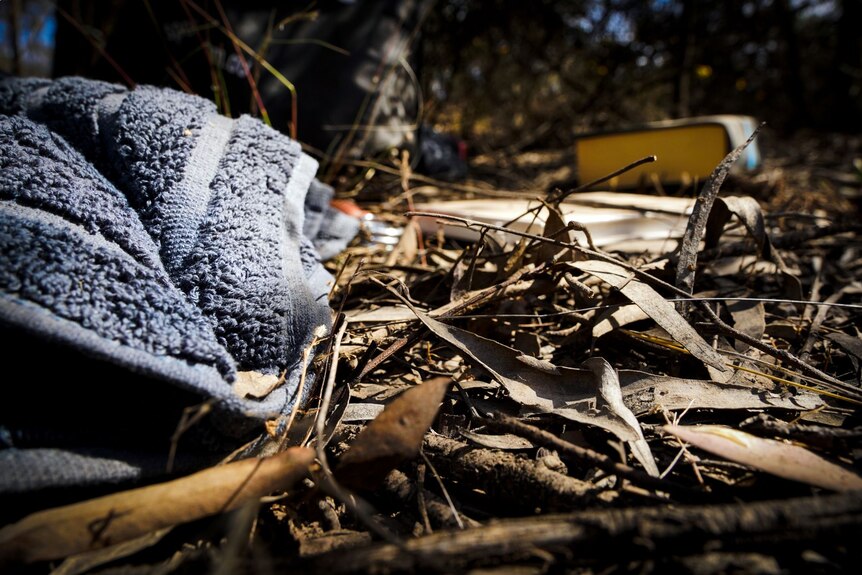 A grey towel lying on the ground in the bush, surrounded by twigs and dried leaves and discarded books.
