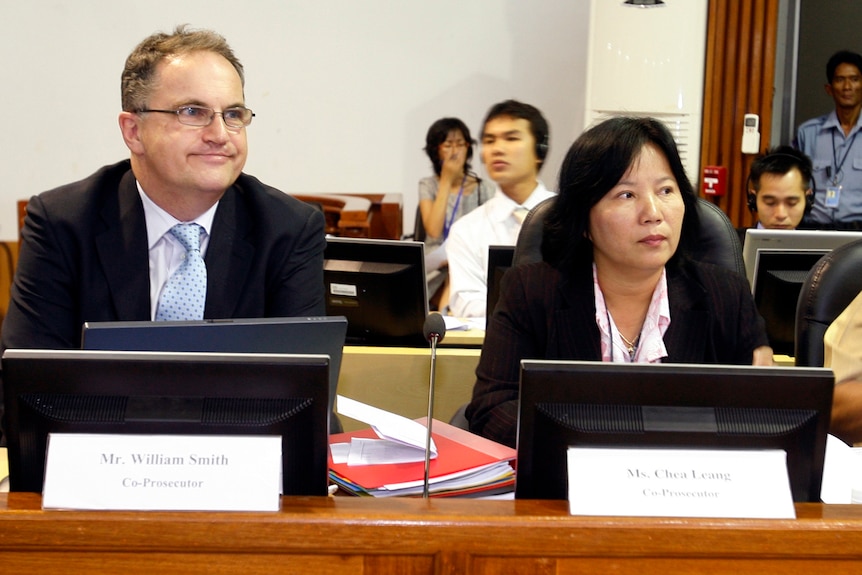 A man and a woman sit at a wooden desk in a courtroom