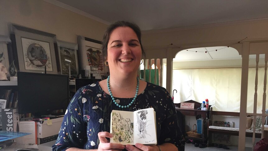 A photo of author in Kathleen Jennings in her house. She is holding a notebook with her drawings in it.