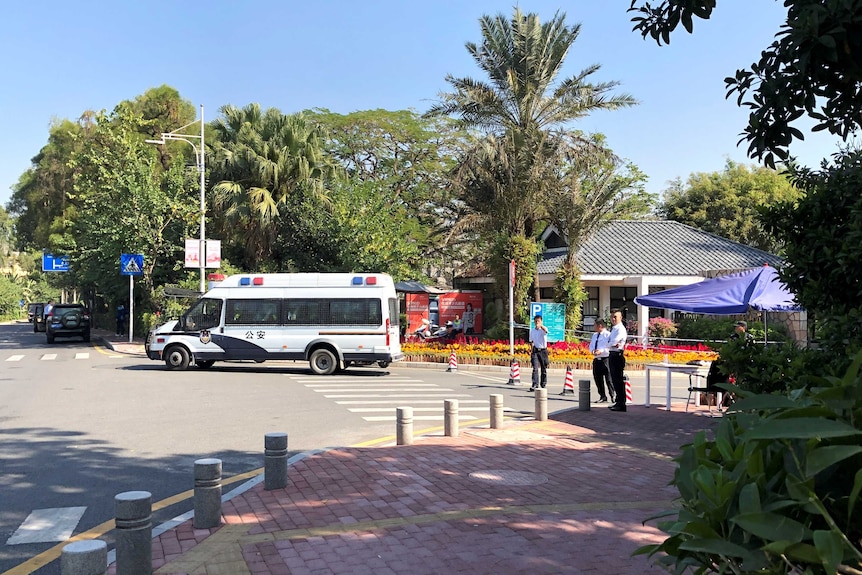 On a clear day, a white Chinese police van leaves a driveway with three security officers monitoring the driveway's entrance.