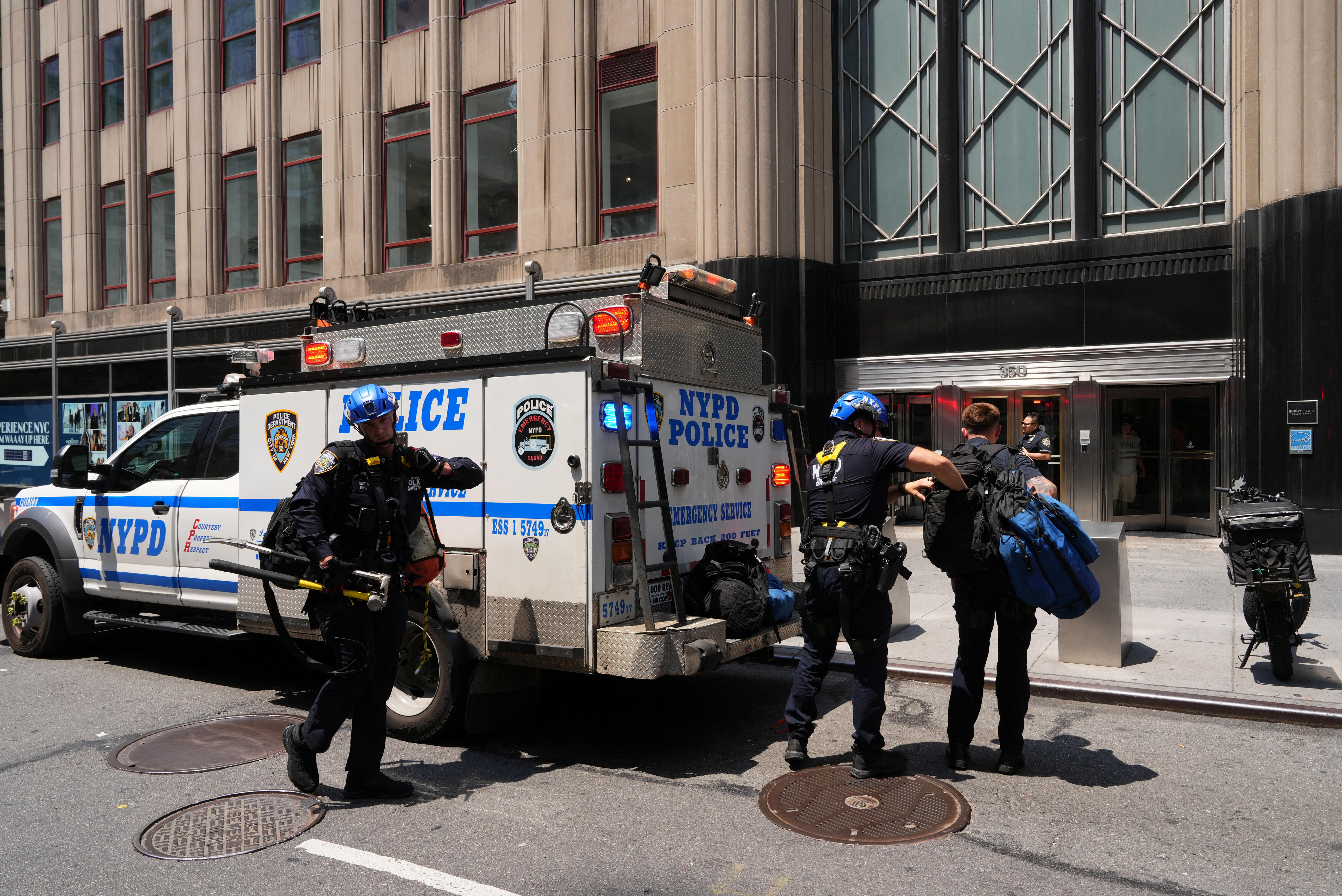 NYPD police officers in protective gear and helmets next to a white and blue patrol car parked on a street.