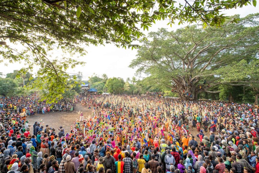 Thousands of colourfully dressed people stand in a clearing surrounded by huge trees.