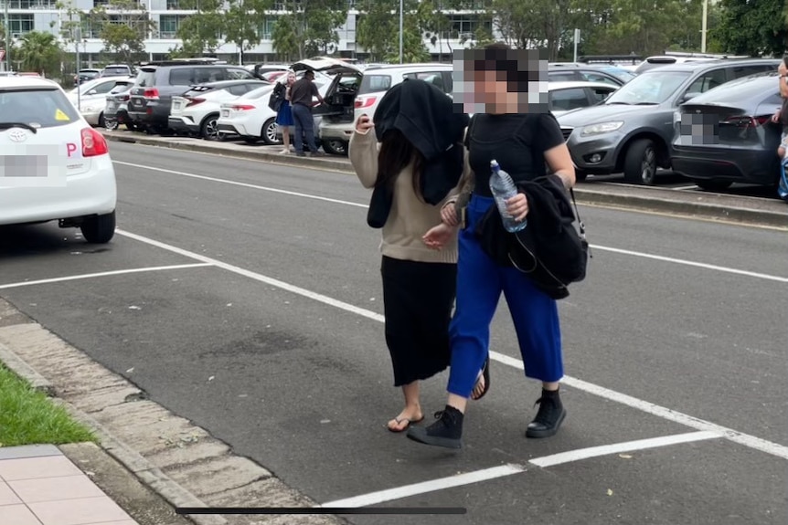 A girl and a woman cross a road together, the girl wearing a black jumper over her head to hide her identify.,