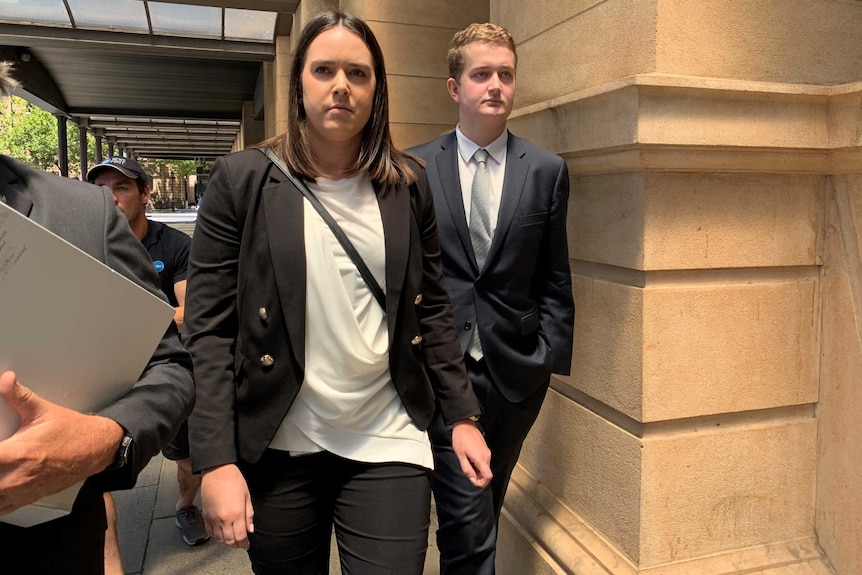 A man in a deep grey suit and white shirt walks alongside a woman in a black suit