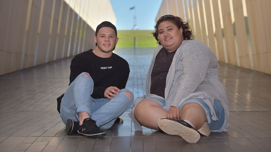 A young man and woman are sitting and smiling at the camera with a blurred corridor in the background