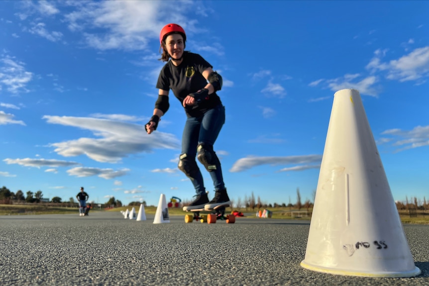 A woman wearing a red helmet and wearing jeans and ankle pads on the slalom skateboard course