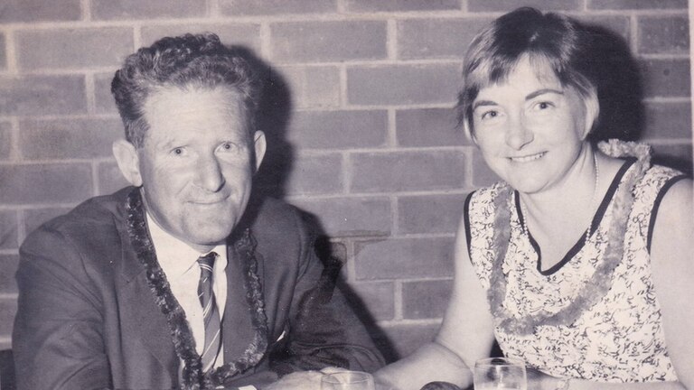 A black and white photo of a man and a woman smiling, sitting at a dinner table with wine glasses, and holding hands.