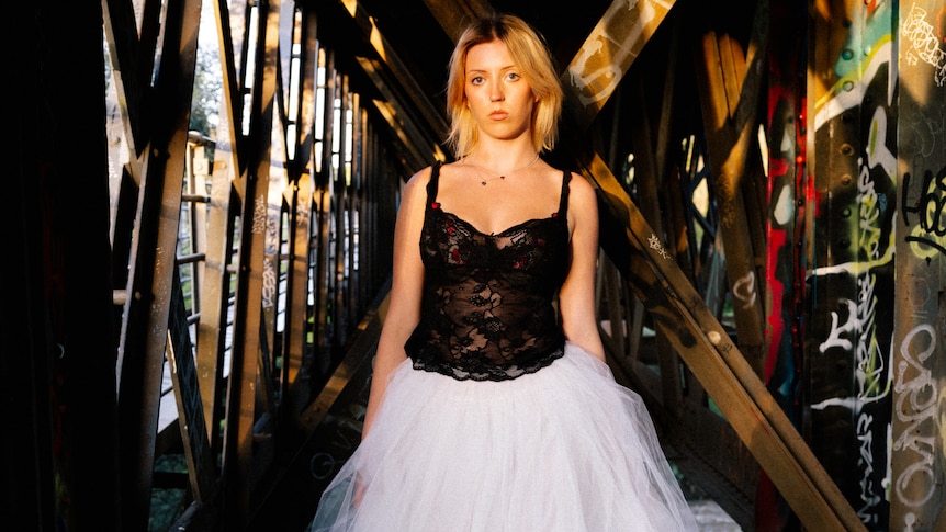 A woman with short blonde hair stands in an industrial space in a big white tulle skirts and a black lace singlet