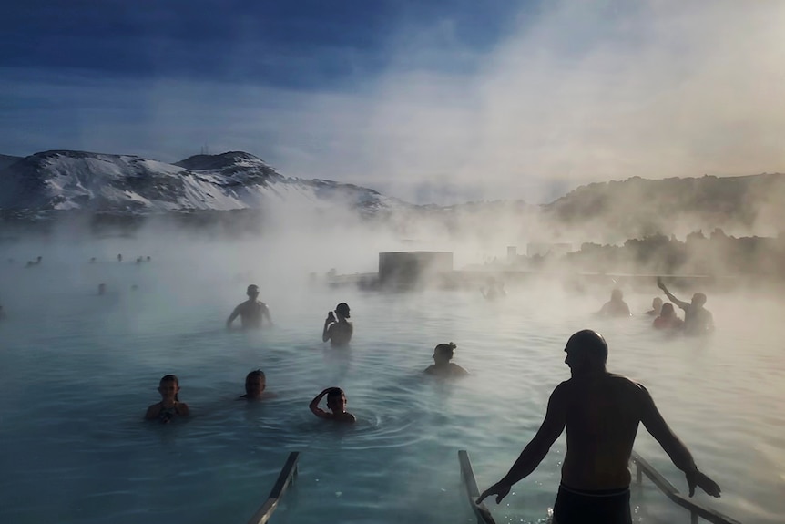 A few dozen people swim in beautiful blue water giving off steam and mist in front of a picturesque mountain range.