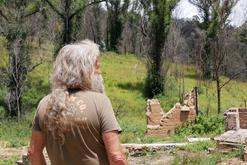 A man with long grey hair stands with his back to the camera looking at the ruins of a brick home set among a green hillside.