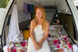 A woman in white dress sits on a bed inside a small van.