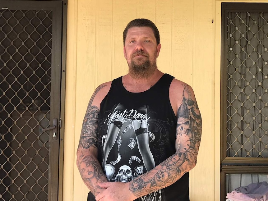 Man with tattoos stands out the front of a yellow house.