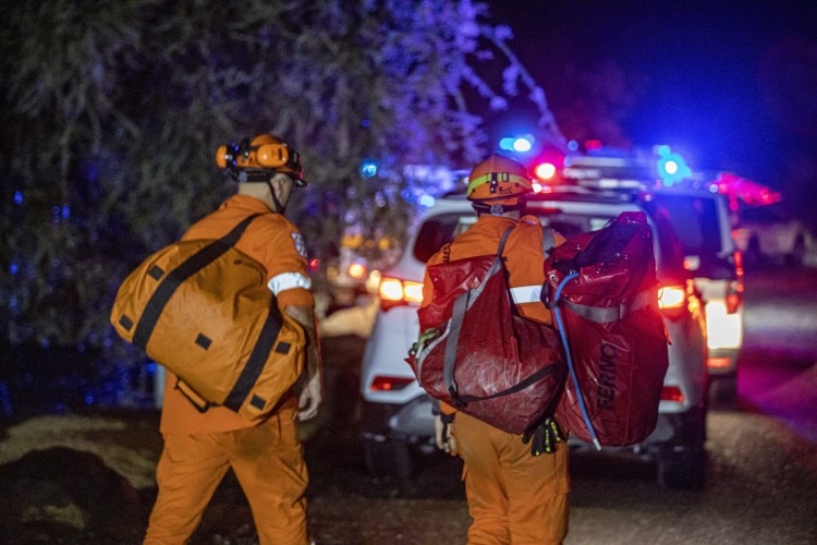 Two firefighters carrying big bags at night, with emergency lights visible in the background.