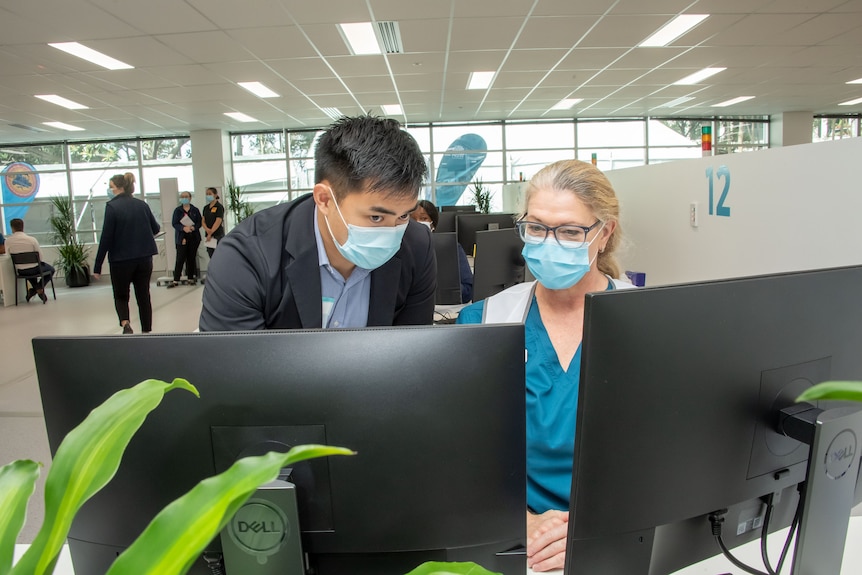 a man wearing a covering over his mouth leans over a woman sitting at a desk in front of a computer