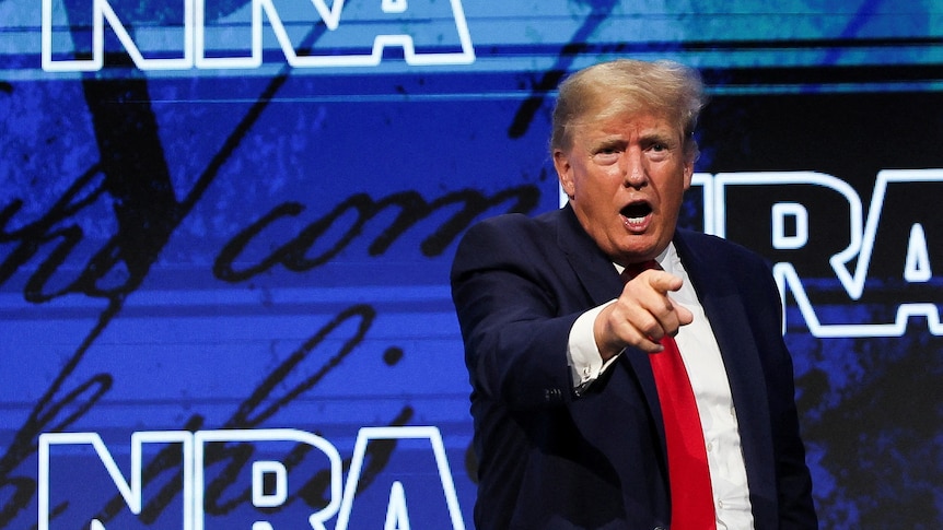 A man in a dark suit points with his right hand as he stands before a blue backdrop reading "NRA".