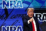 A man in a dark suit points with his right hand as he stands before a blue backdrop reading "NRA".