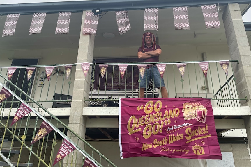 man stands on balcony with go queensland go maroon flag hanging from balcony 