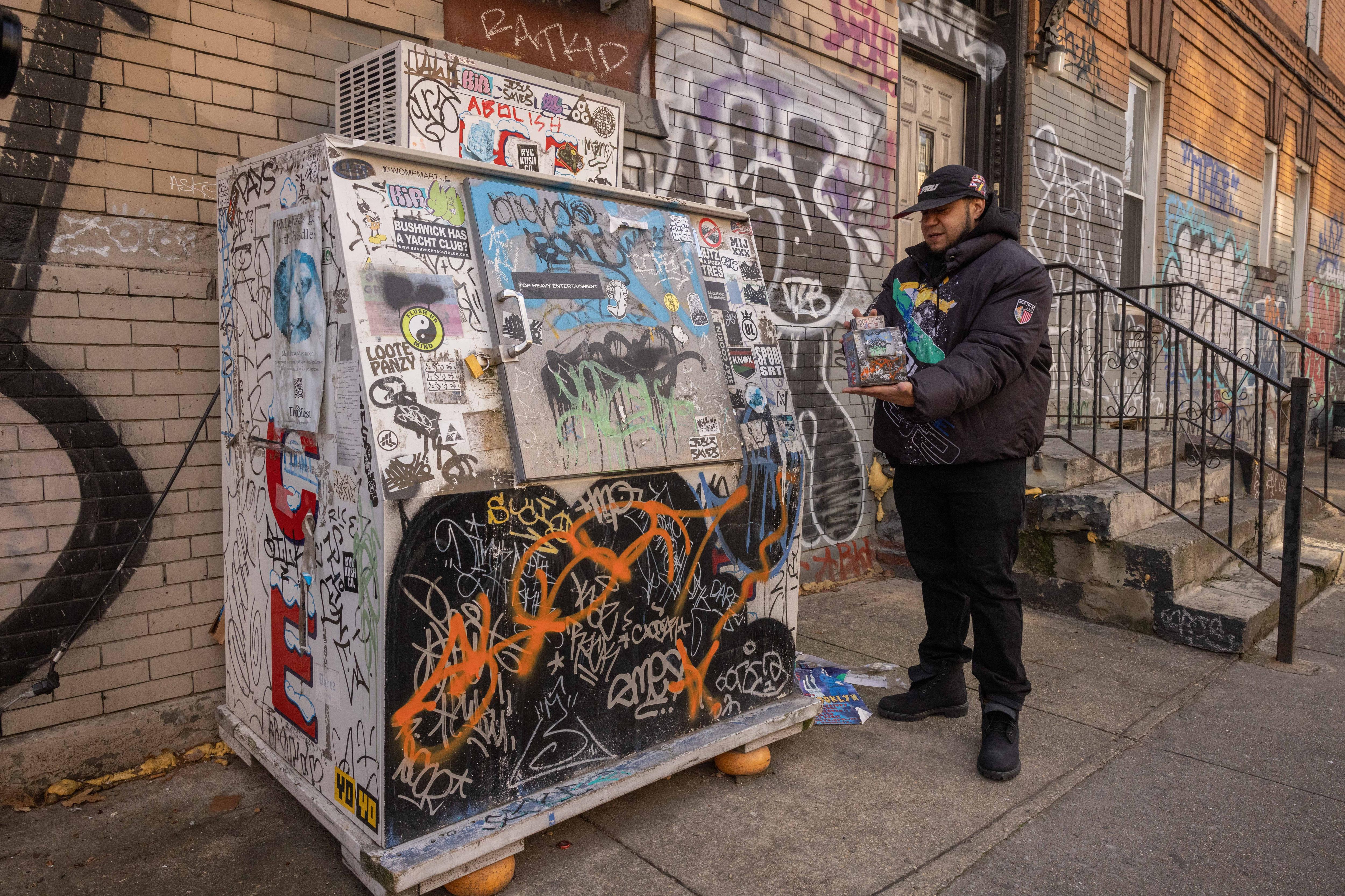 A man dressed in streetwear stands next to a commercial ice box holding a minature version of the same thing in his hands