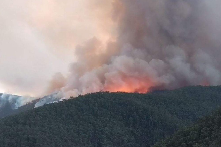 An orange glow and smoke can be seen over the crest of a series of mountains