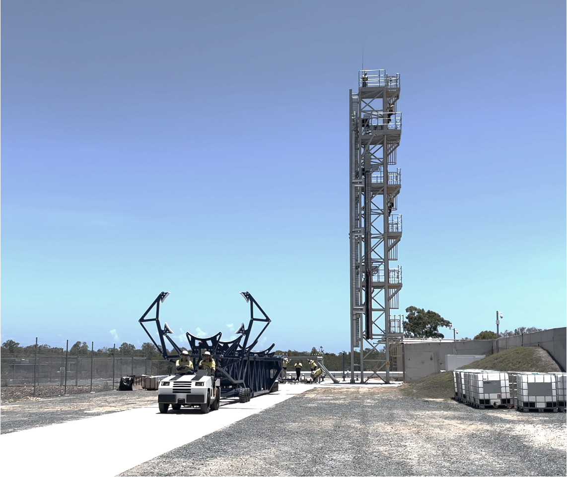 A ute carries a rocket holder in the foreground with a rocket launch tower in the background