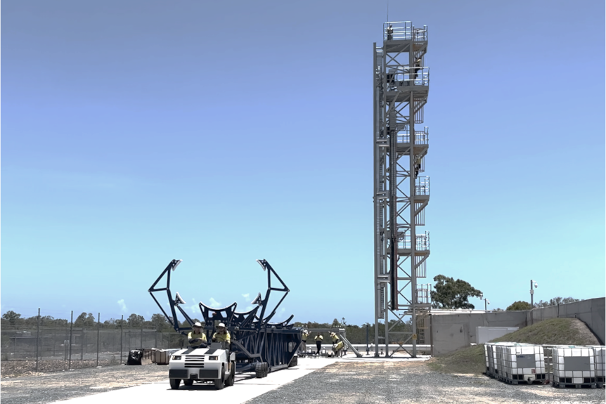 A ute carries a rocket holder in the foreground with a rocket launch tower in the background