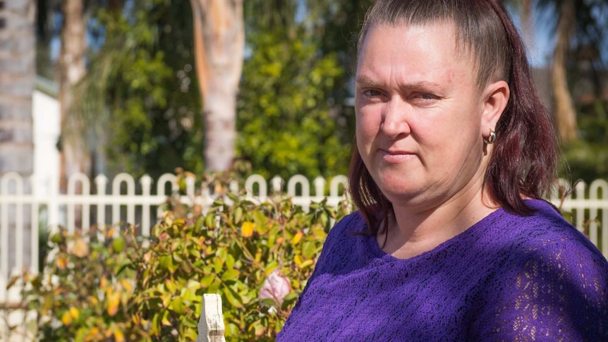 A woman in a purple top stands by the white picket garden gate of a country home.