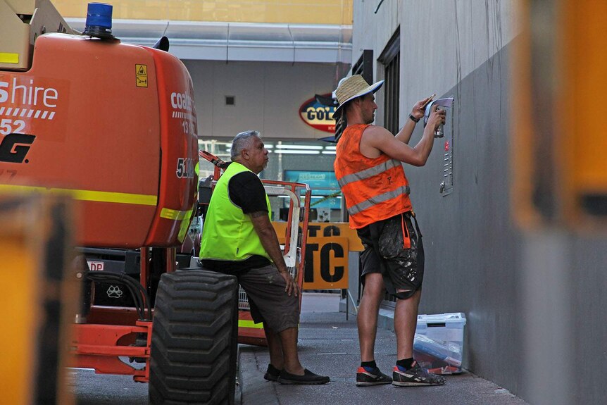 Two artists look at the wall they will paint over the next week. One of them sits on a cherry-picker.