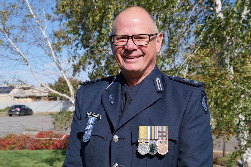A police officer in blue jacket with medals and pins with no hair, black glasses stands in front of a tree