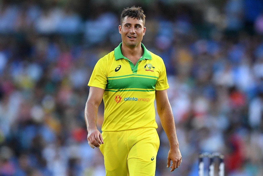 A man wearing a yellow shirt with a green stripe walks towards the camera with a grin on his face