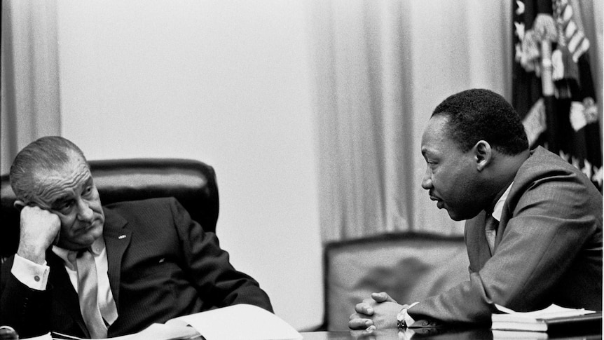 Martin Luther King and President Johnson sit at a table. The president looks unamused as King talks. Black and White.