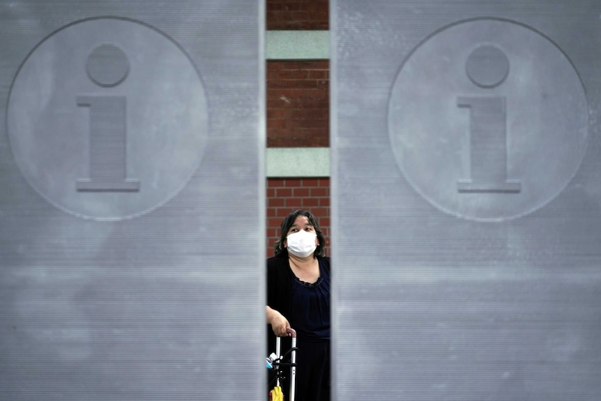 A woman stands wearing a white face mask and holding the handle of a suitcase