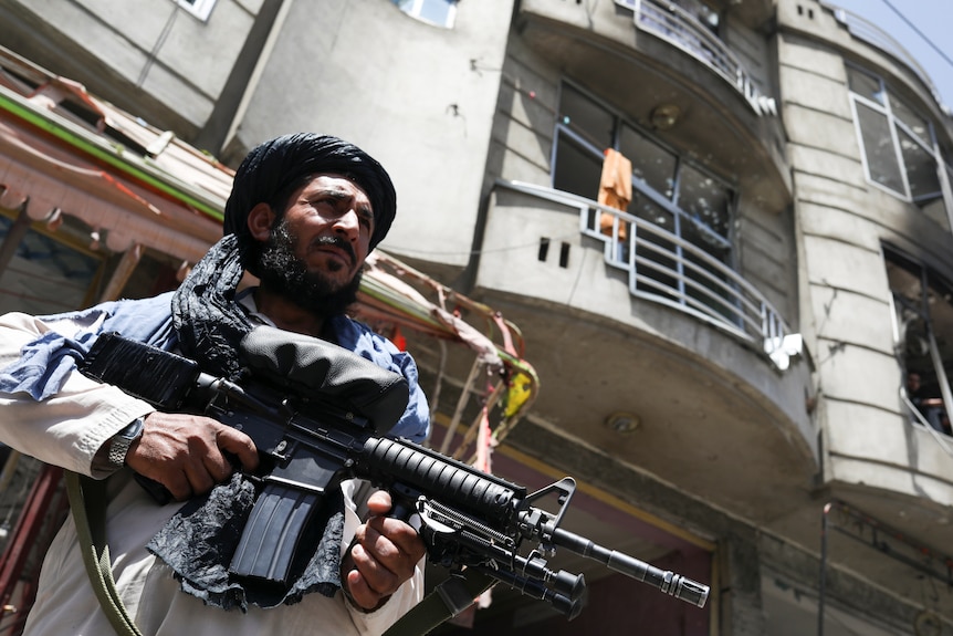 A man carrying an automatic weapon stands in front of a building that appears to have been damaged by an explosion