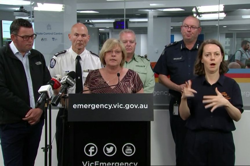 Lisa Neville stands at a lectern speaking to the media, flanked by Premier Daniel Andrews and emergency services leaders.