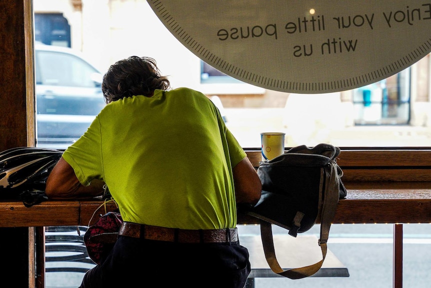 A person with a bright t-shirt sits leaning over a cafe-style bench next to a window overlooking a street with a car.