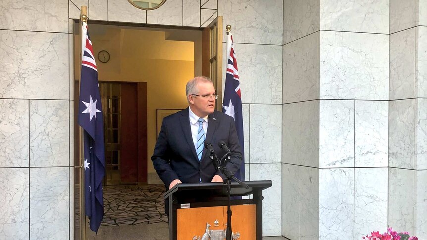 Scott Morrison wearing a pale blue tie stands behind a lectern in front of two Australian flags.